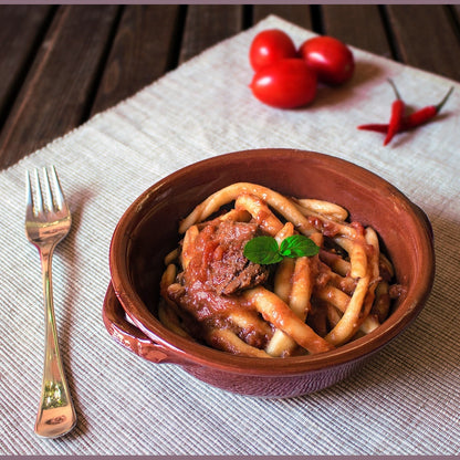 Ragù di maiale pronto alla calabrese servito con pasta in una ciotola di terracotta, decorato con pomodorini e peperoncino.